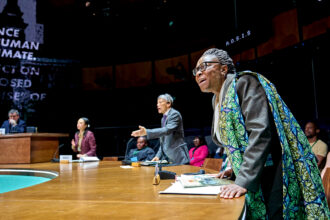 Aïcha Kossoko as Tanzania, Kwong Loke as China, and Andrea Gatchalian as Kiribati standing at their seats and leaning forward in objection.