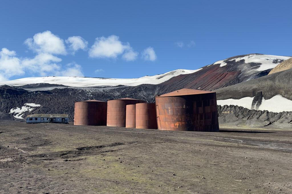In Whalers Bay, relics of a bygone era of whale hunting remain. Whale bones poke through ashen sediment near dilapidated wooden boats whalers used to tow the animals to shore where their blubber was harvested to produce lamp oil. The oil was then stored in tall metal vats before exporting it. Credit: Teresa Tomassoni/Inside Climate News