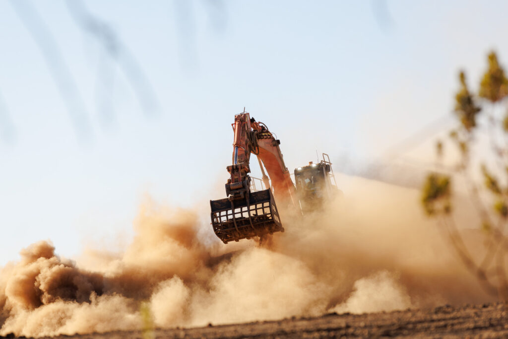 An excavator moves dirt on a construction site, creating a large cloud of dust under a clear sky.