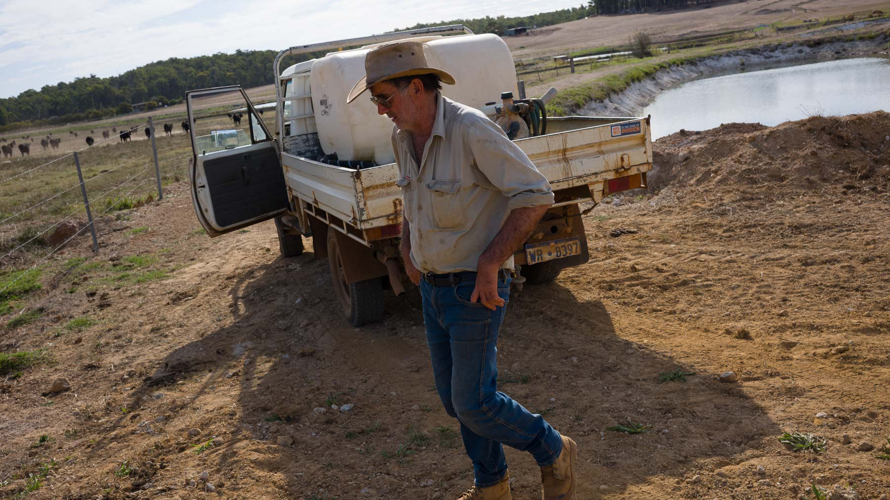 A man in work clothes and a hat walks beside a utility truck parked near a pond on a rural property, with fields and trees in the background.