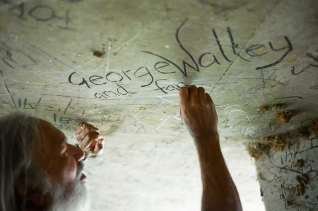 An older man writes "George Walley and family" on a graffiti-covered concrete surface with a charcoal.