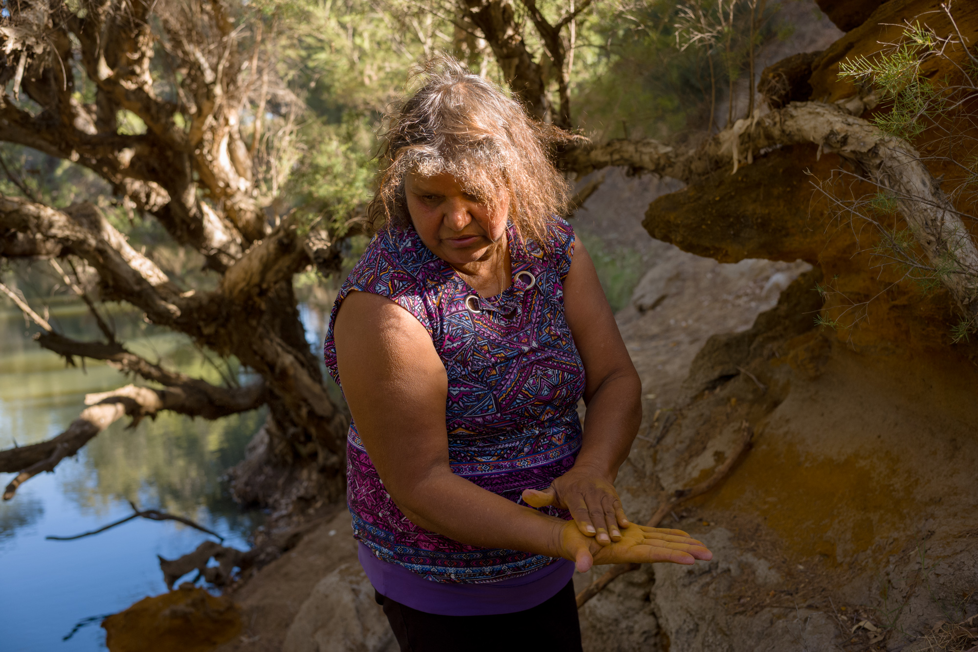 A woman stands by a riverbank, rubbing red dirt on her hand, with trees and water in the background.