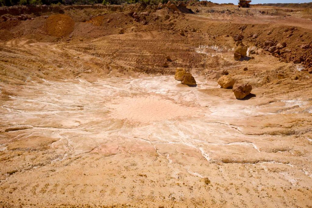Wide view of a dry, cracked earth crater with scattered boulders and reddish-brown soil, under clear daylight.