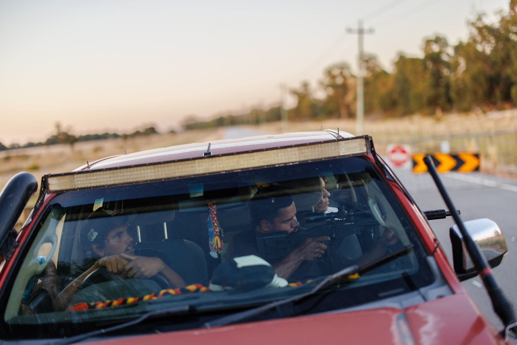 Three people sit inside a red vehicle; one person in the passenger seat is aiming a rifle out the window, while the driver looks forward. Road signs and trees are visible outside.
