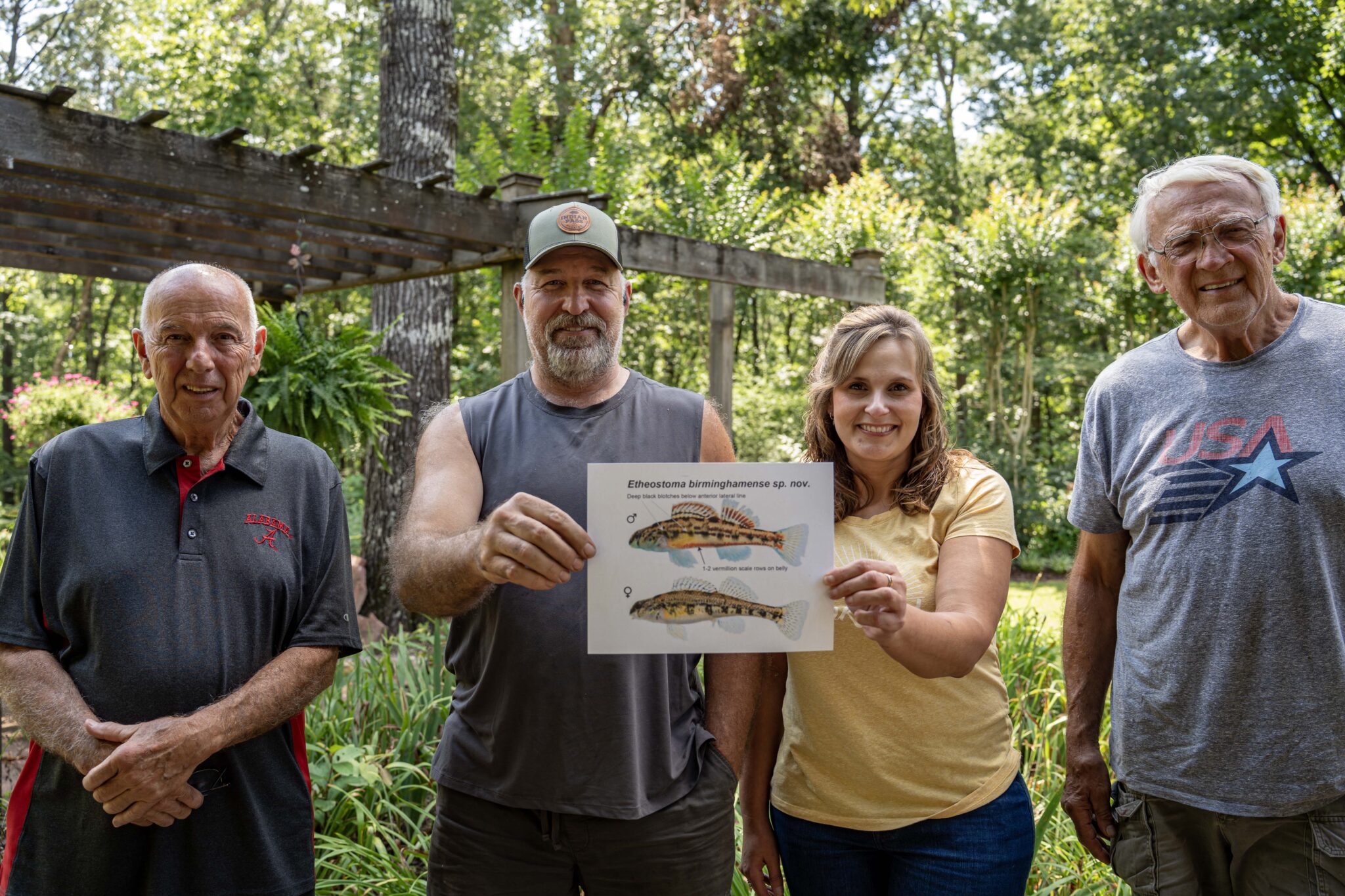 Four people are standing out of doors, trees in the background. The two in the middle are holding a piece of paper with the colorful Birmingham darter.