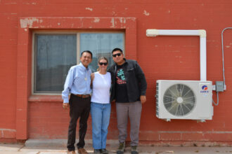 Red Feather staff (from left) Duane Tsinigine, Tavanne Sousa and Tyler Puente stand next to a heat pump installed with the nonprofit’s help at a home on the Hopi Reservation. Credit: Wyatt Myskow/Inside Climate News