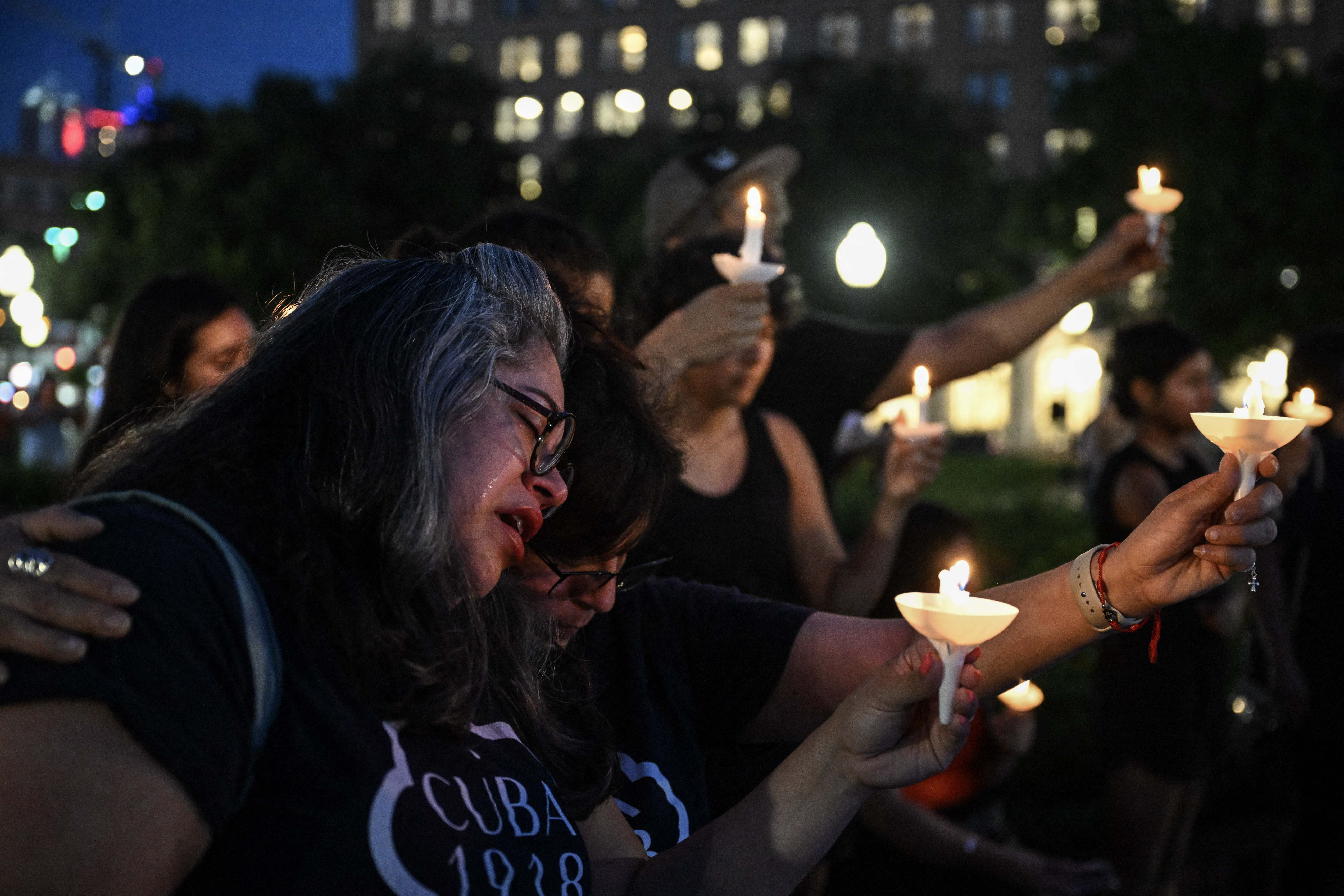 People hold candles in a park. In the foreground, a woman weeps.