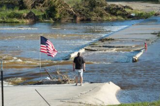 A man stands with his back to the camera near an American flag at the bank of the river, looking at the damage