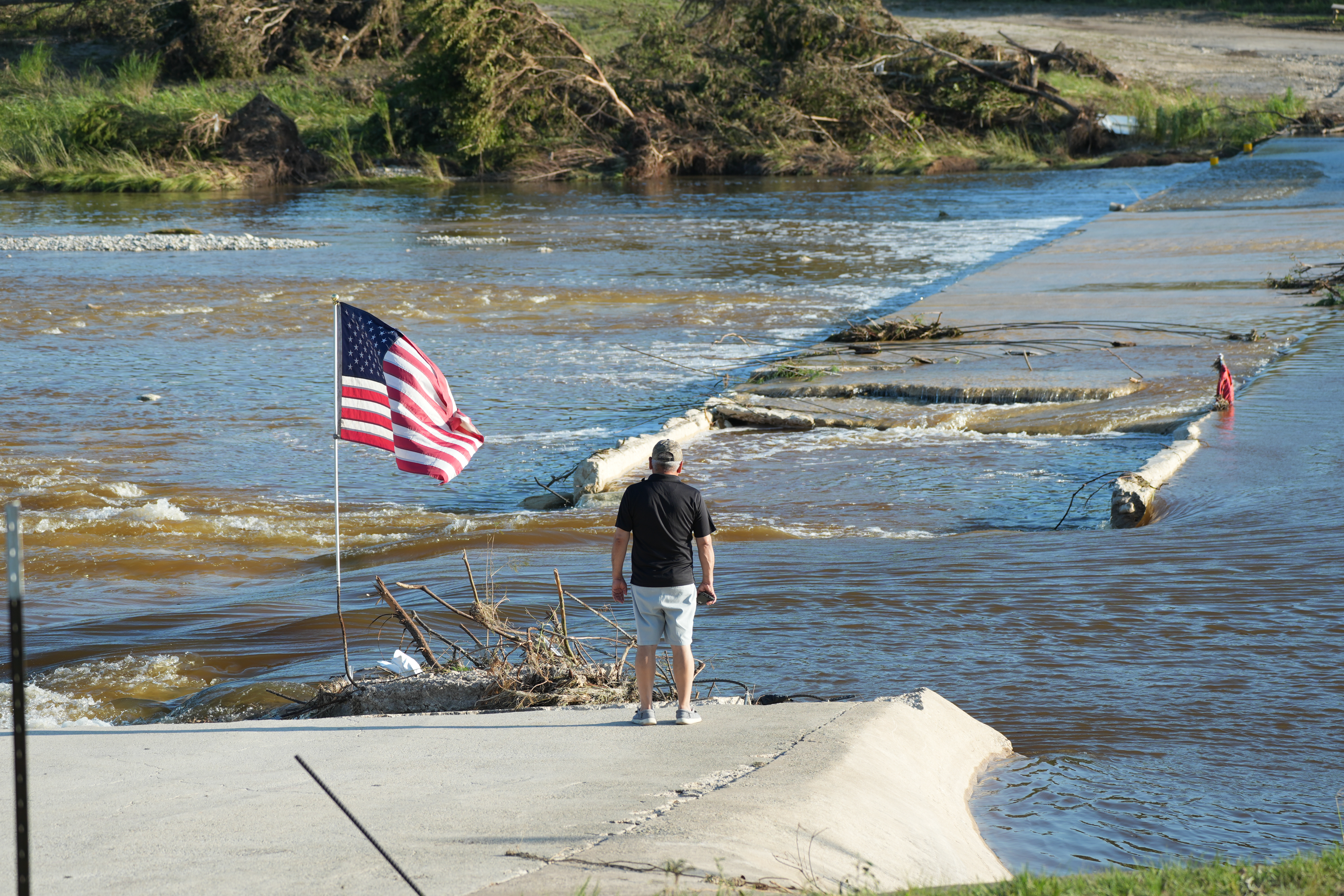 A man stands with his back to the camera near an American flag at the bank of the river, looking at the damage