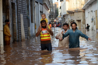 A child sits on the shoulders of a rescue worker, who is wading through the water near other people. The water is reaching the adults' upper thighs. Buildings rise on either side of them.
