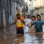 A child sits on the shoulders of a rescue worker, who is wading through the water near other people. The water is reaching the adults' upper thighs. Buildings rise on either side of them.