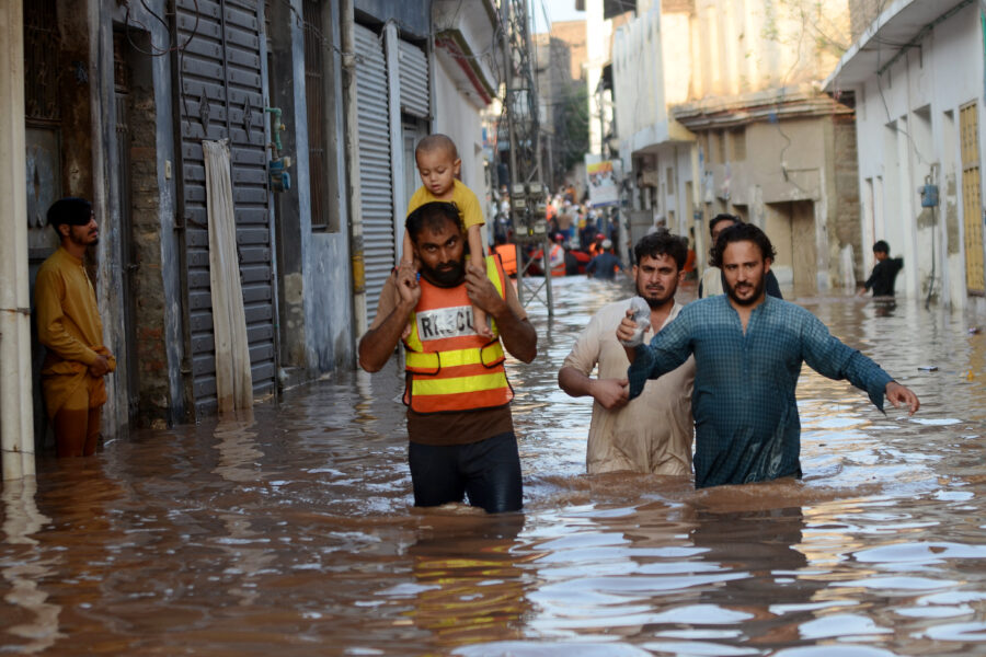 A child sits on the shoulders of a rescue worker, who is wading through the water near other people. The water is reaching the adults' upper thighs. Buildings rise on either side of them.