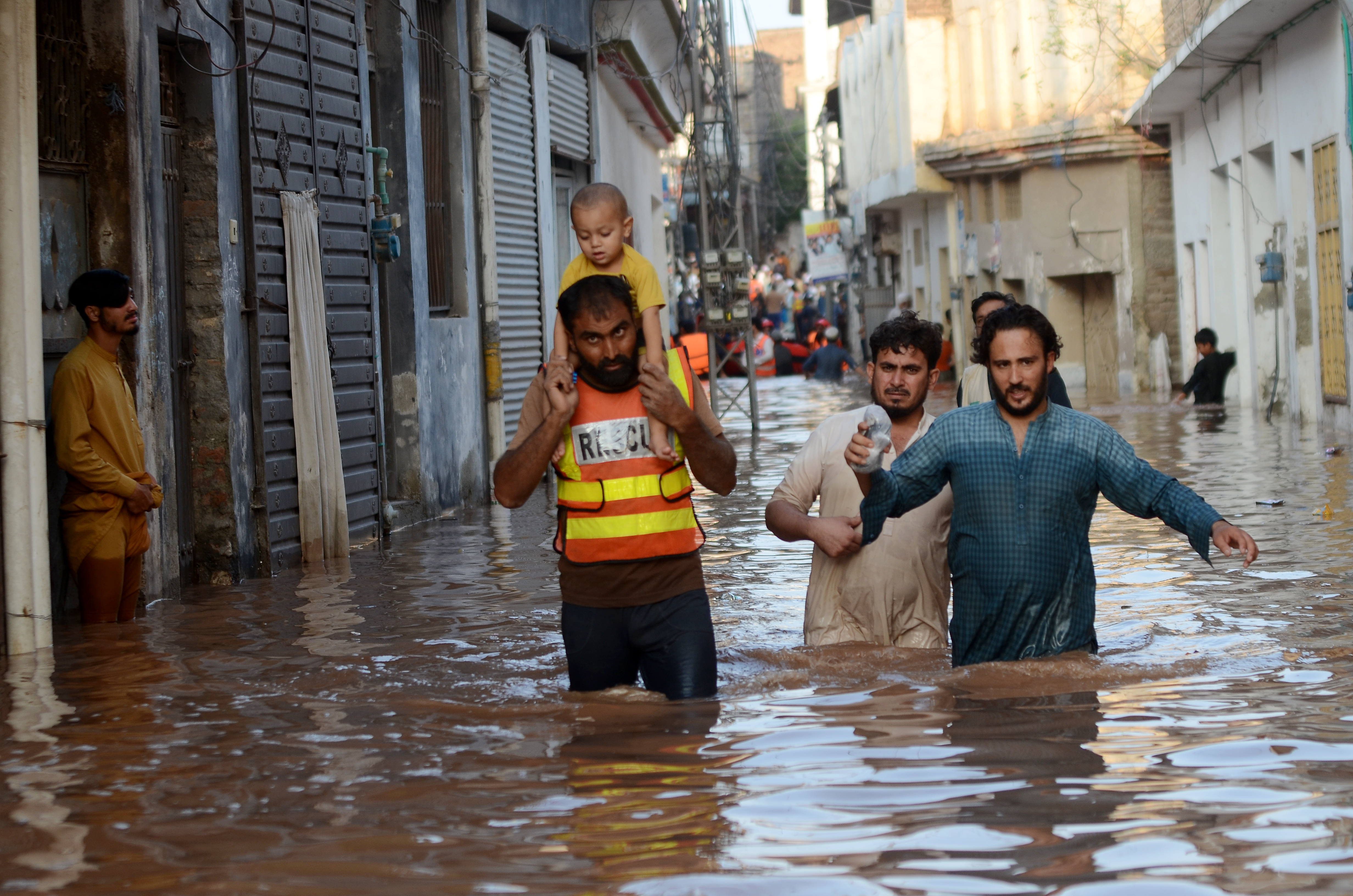 A child sits on the shoulders of a rescue worker, who is wading through the water near other people. The water is reaching the adults' upper thighs. Buildings rise on either side of them.