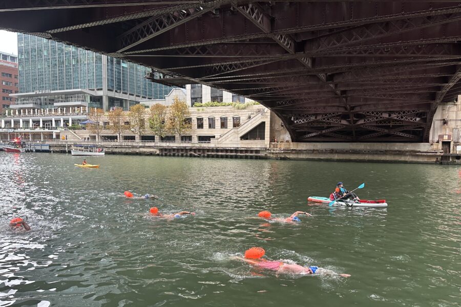 People in blue swim caps and neon orange buoys swim under a bridge in the Chicago River past people in kayaks