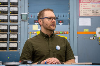 Nicholas Spada stands in front of an instument panel at UC Davis’ Crocker Nuclear Laboratory with a radiation exposure monitor prominently pinned to his shirt’s pocket on March 25.