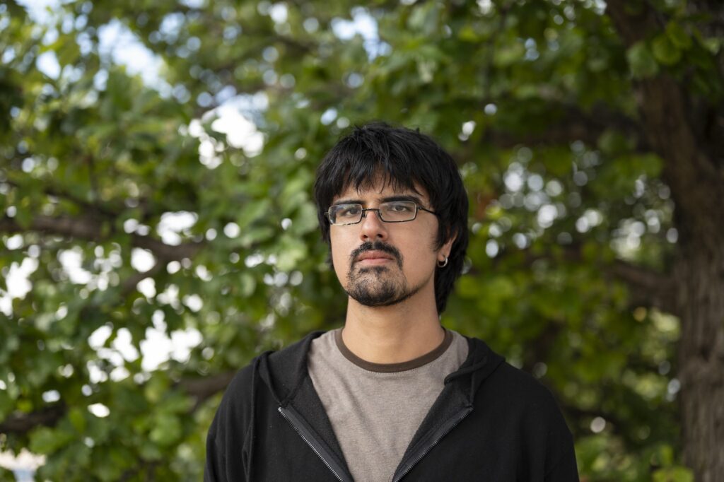 Ajay Chatha looks into the camera with a serious expression. Beyond is the out-of-focus greenery of tree leaves.
