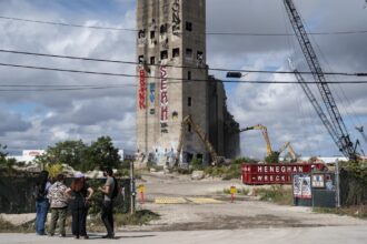 The organizers are standing in a cluster in the foreground. Beyond them are the silos, tagged with graffiti, and demolition equipment.