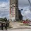 The organizers are standing in a cluster in the foreground. Beyond them are the silos, tagged with graffiti, and demolition equipment.