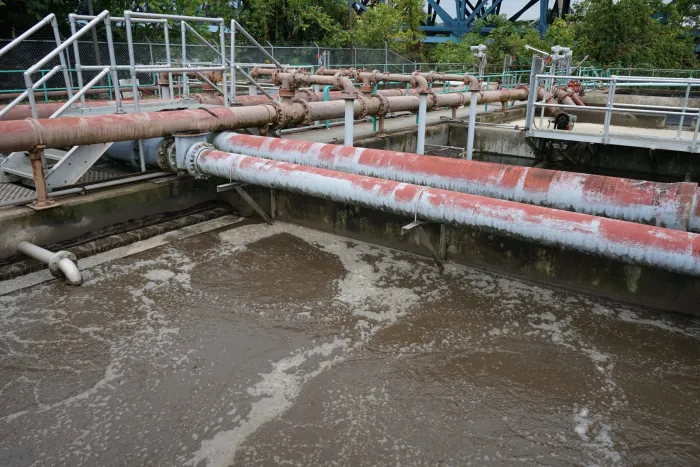 A number of pipes and liquid beneath them are seen in the foreground, with trees in the background