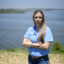 Jackie Medcalf is looking at the camera with a serious expression, arms crossed. Behind her is the river.