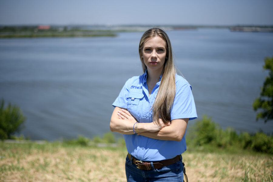 Jackie Medcalf is looking at the camera with a serious expression, arms crossed. Behind her is the river.