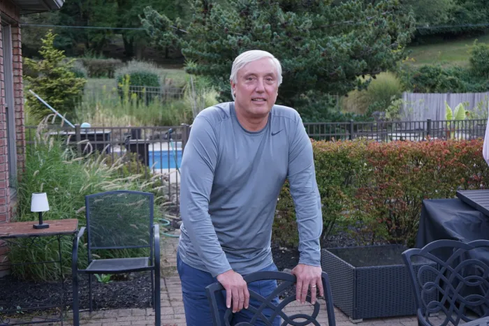 James Sepesky is standing in his yard, holding the back of a metal chair and looking into the camera.