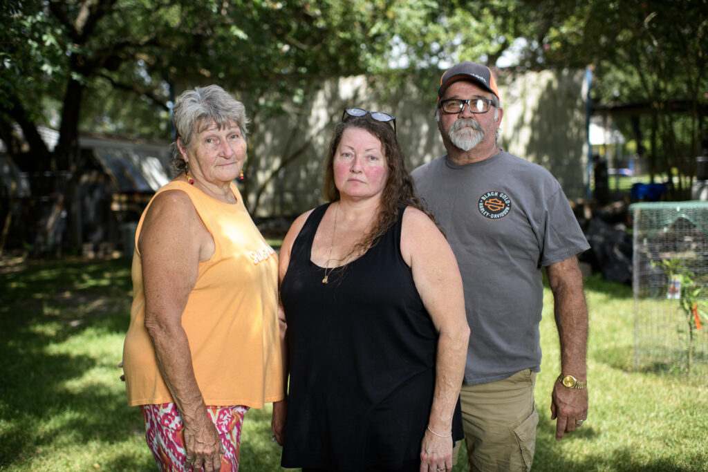 All three are standing outside, grass and trees behind them