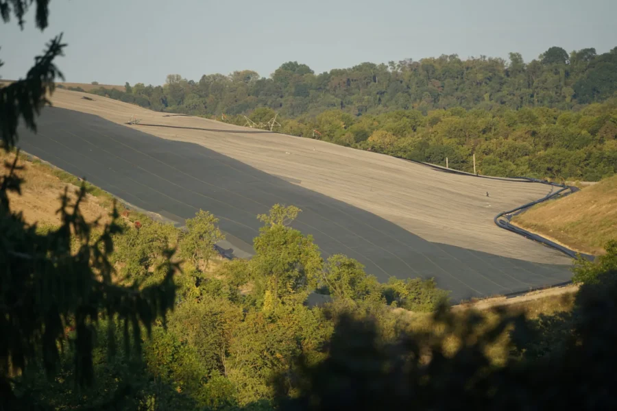 A landscape of the landfill appears in the middle ground, a brownish-sandy color, with trees at either side.