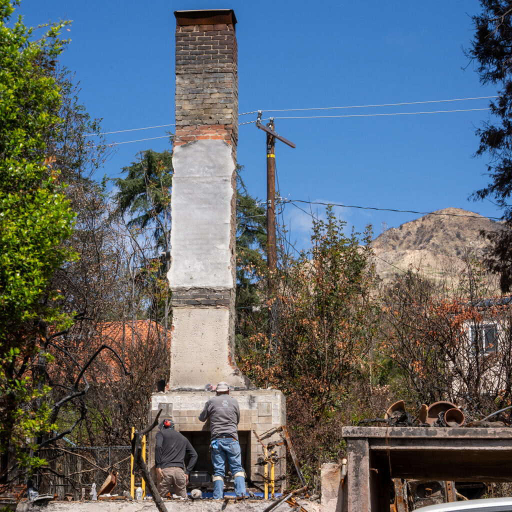 Crews work to clear rubble and disassemble the burned husk of the fireplace and chimney where the house across the street once stood.