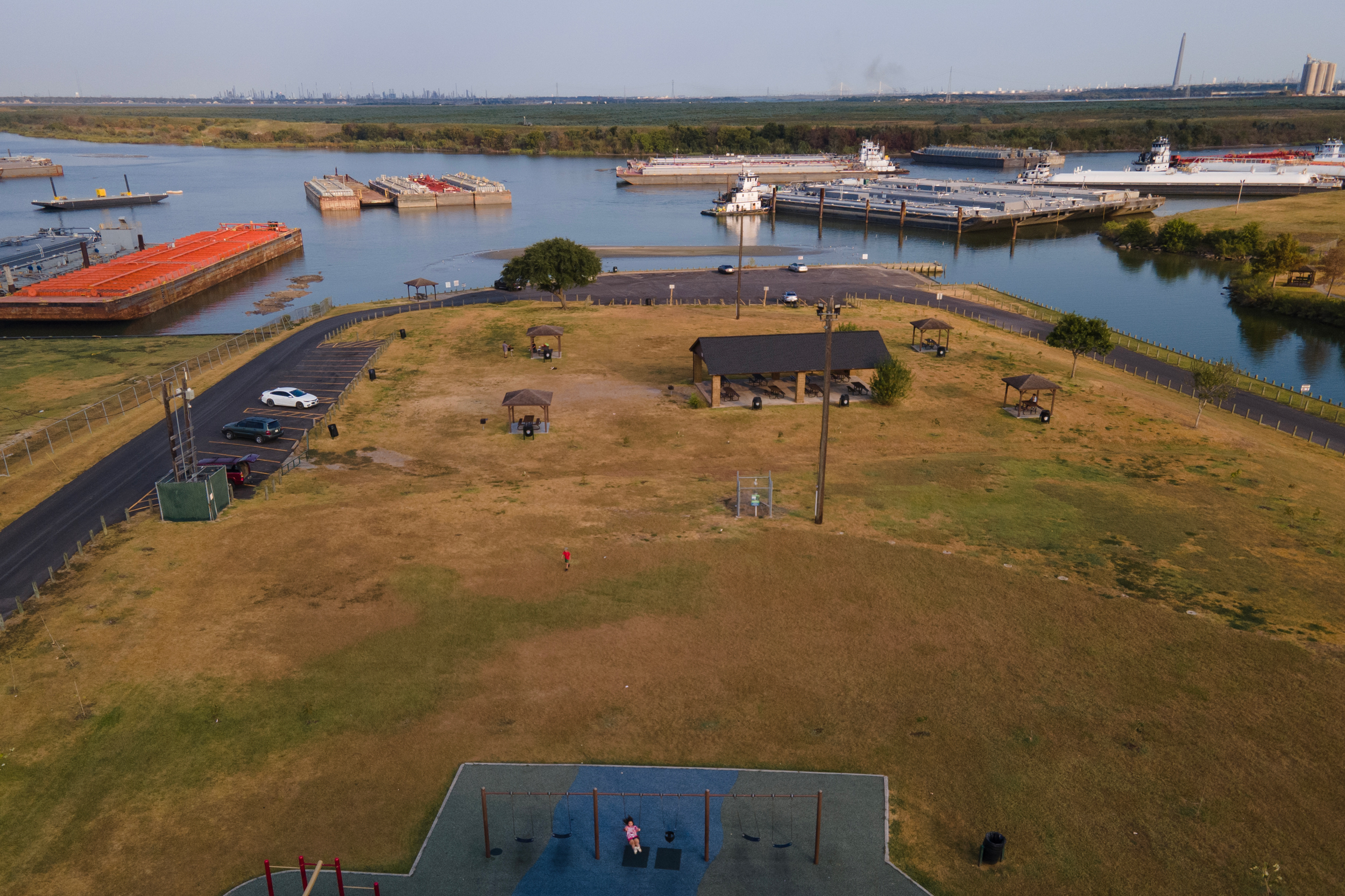 An aerial photo shows a child on a swingset in the foreground. Beyond that is the rest of the park, including a small parking lot, and the barges on the river.