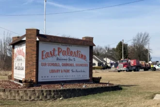 View shows the East Palestine welcome sign. Homes and a truck are visible behind it.