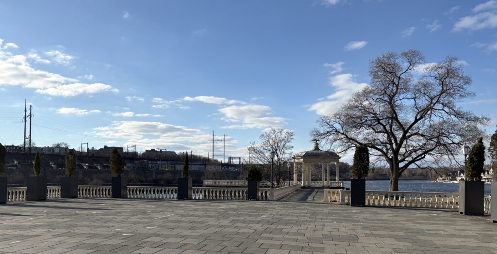 The view is from the edge of the Water Works property overlooking the river. At the waterside, there is a fence, several trees and a small gazebo.
