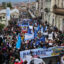 An aerial photo shows a large crowd filling a street. Some people hold signs or banners.