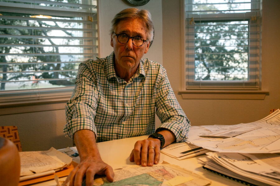 James Dodson is looking at the camera with a serious expression. On the table in front of him are maps and documents. Behind him are windows, the shades open, trees beyond them.