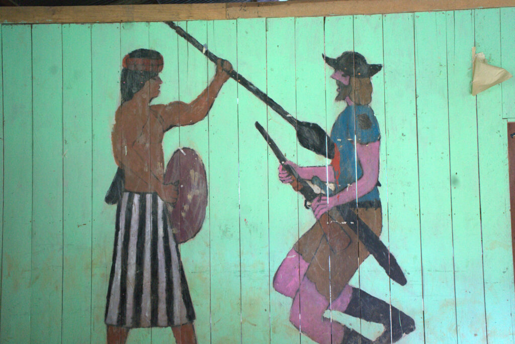 A mural in the Maikiuants community center depicts an Indigenous warrior spearing a colonist.