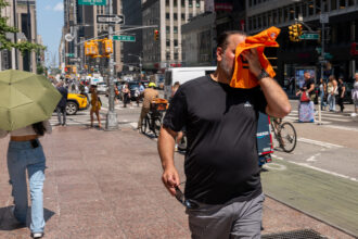 A man on the street wipes his face with orange fabric and another pedestrian holds an umbrella to block out the sun.