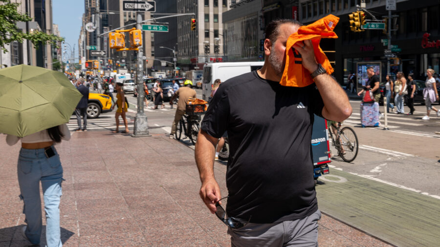 A man on the street wipes his face with orange fabric and another pedestrian holds an umbrella to block out the sun.