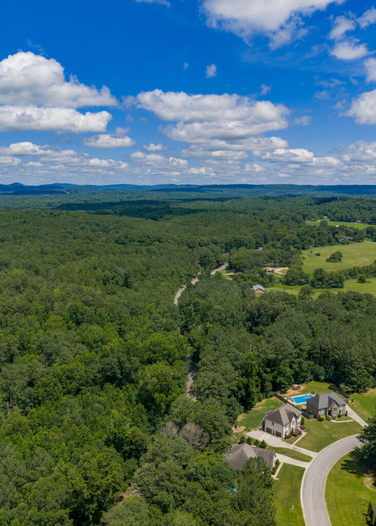 An aerial shot shows a blue sky with white clouds, a swath of forest and a residential cul-de-sac with several houses.