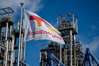 A TotalEnergies flag flies in the foreground, with a refinery in the background.