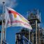 A TotalEnergies flag flies in the foreground, with a refinery in the background.