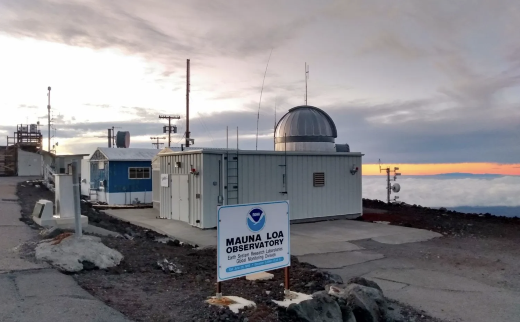 A few small buildings with scientific equipment are shown in an exterior view with a sky lit by either sunrise or sunset and a sign that says, "Mauna Loa Observatory" with a NOAA logo