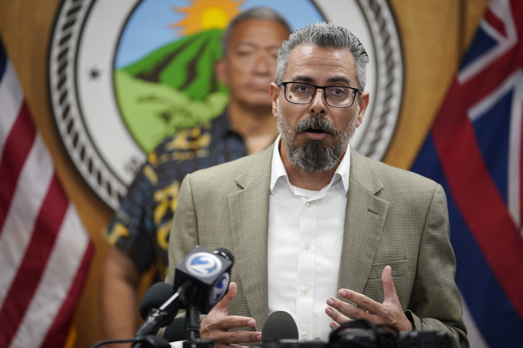 Maui Prosecutor Andrew Martin is pictured speaking into television microphones from a podium during a 2023 press conference after the wildfires
