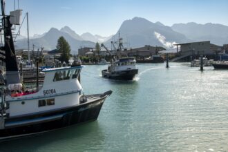 A commercial fishing boat enters the harbor at Valdez, Alaska, with a view of the Chugach mountain range.