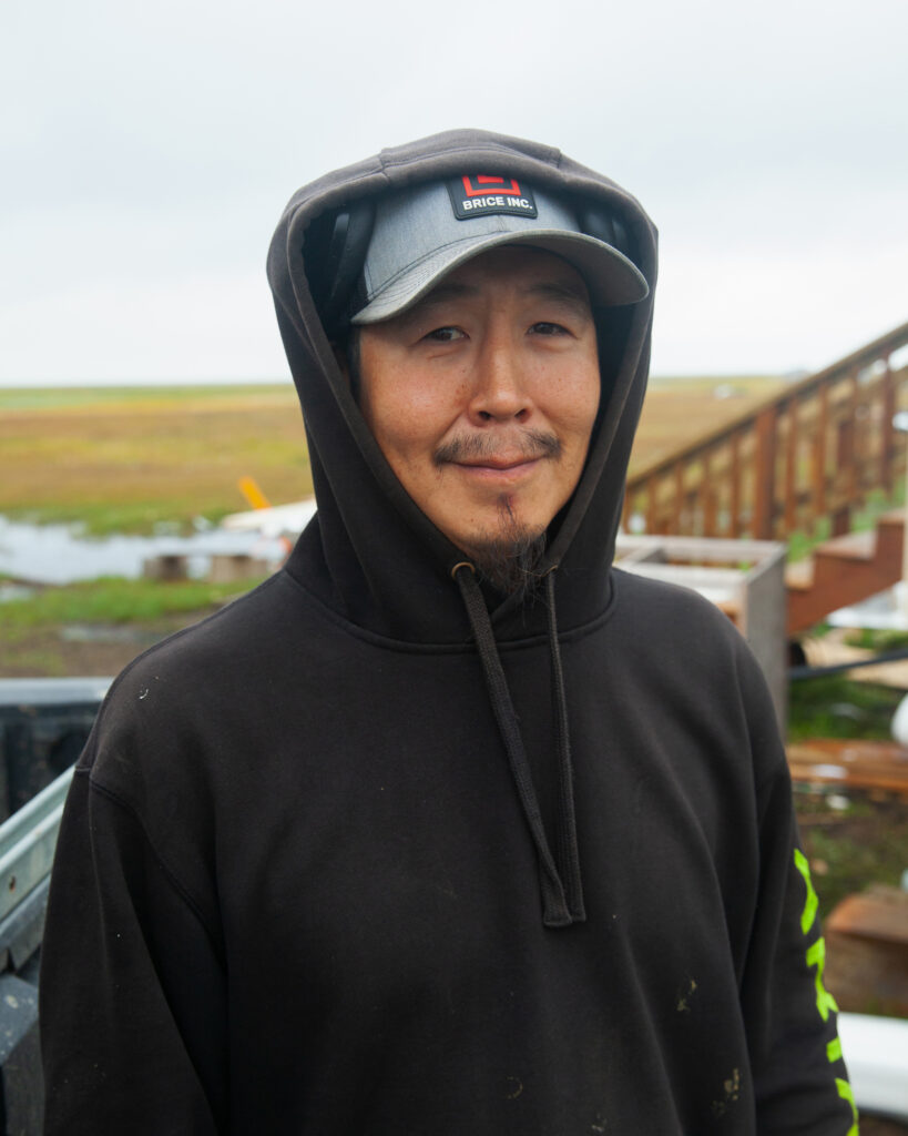 Lewis Amik III stands in front of his damaged house in Kwigillingok. He came back to the village three weeks after the typhoon to “keep the lights on” at the local power plant where he works. Credit: Olga Loginova/Inside Climate News
